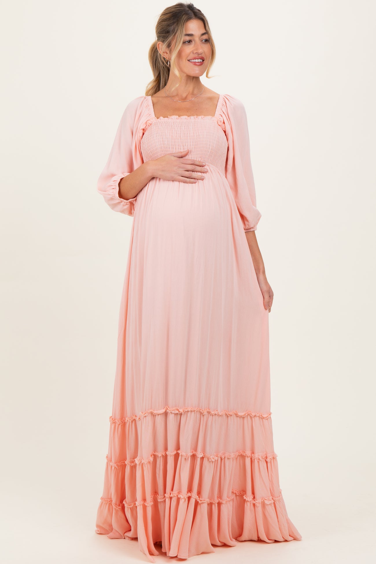 woman wearing a light peach maternity maxi dress with smocked bodice and tiered ruffle hem, holding her baby bump