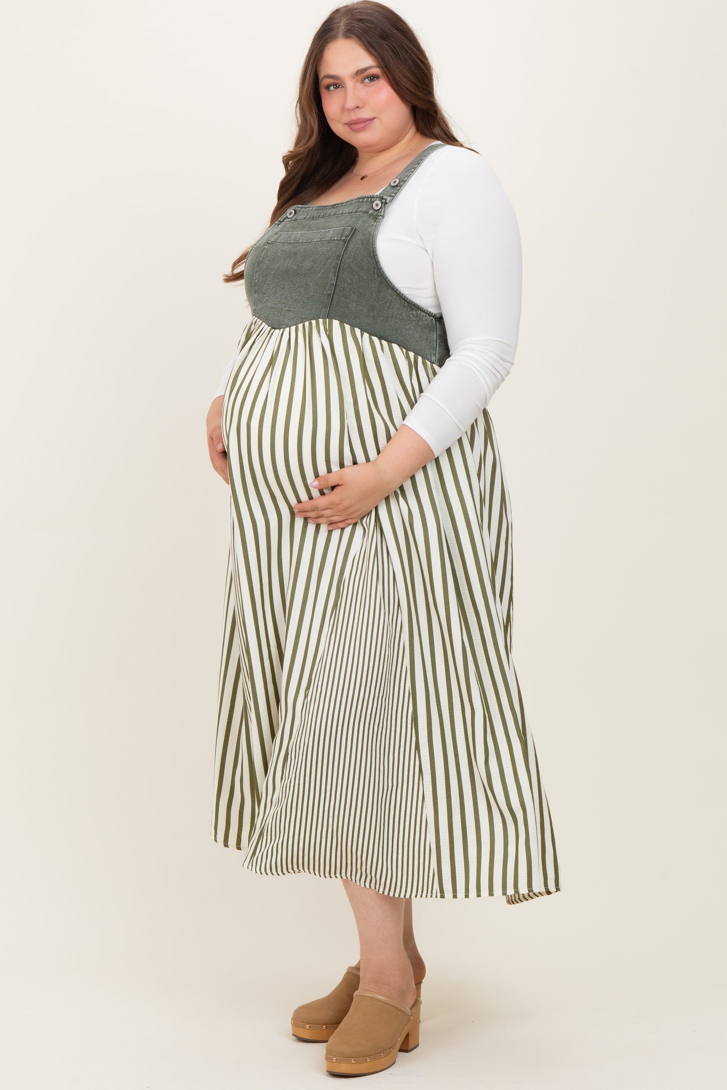 woman wearing a green and white striped maternity dress with overalls, holding her baby bump