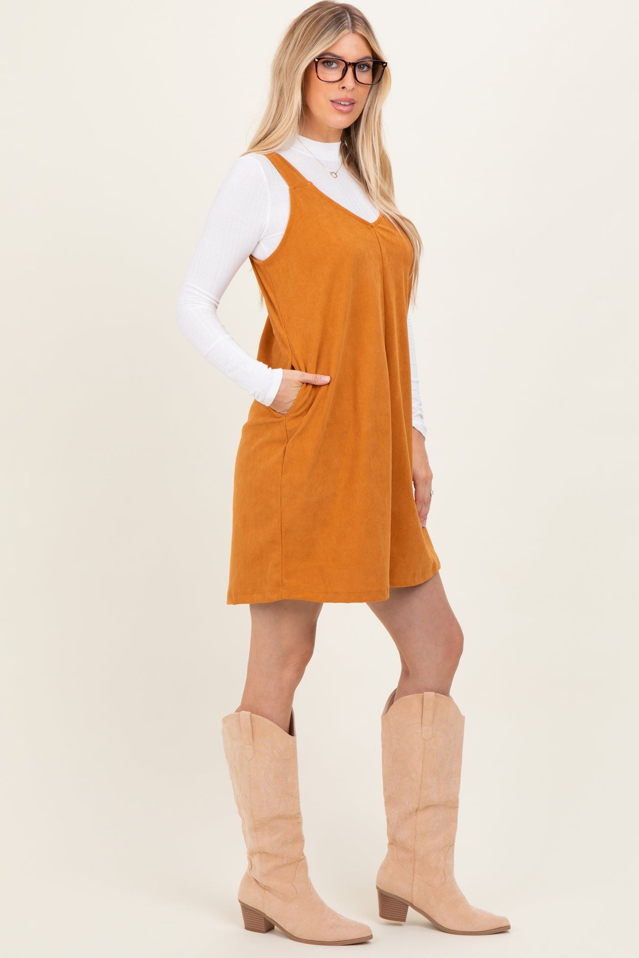 woman wearing an amber suede sleeveless dress with pockets and a white long sleeve top, paired with beige knee-high boots