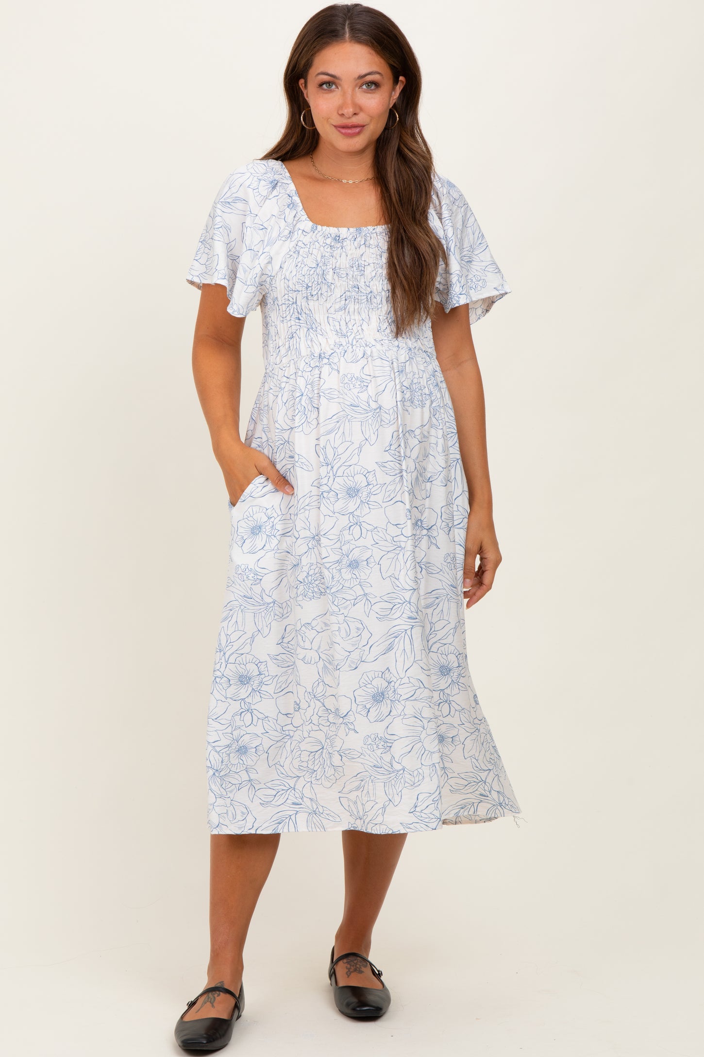 woman wearing a white floral print maternity dress with short sleeves and pockets, standing against a neutral background