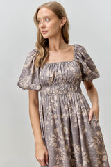 woman wearing a brown floral puff sleeve dress with a square neckline and elastic waist, posing with hand in pocket