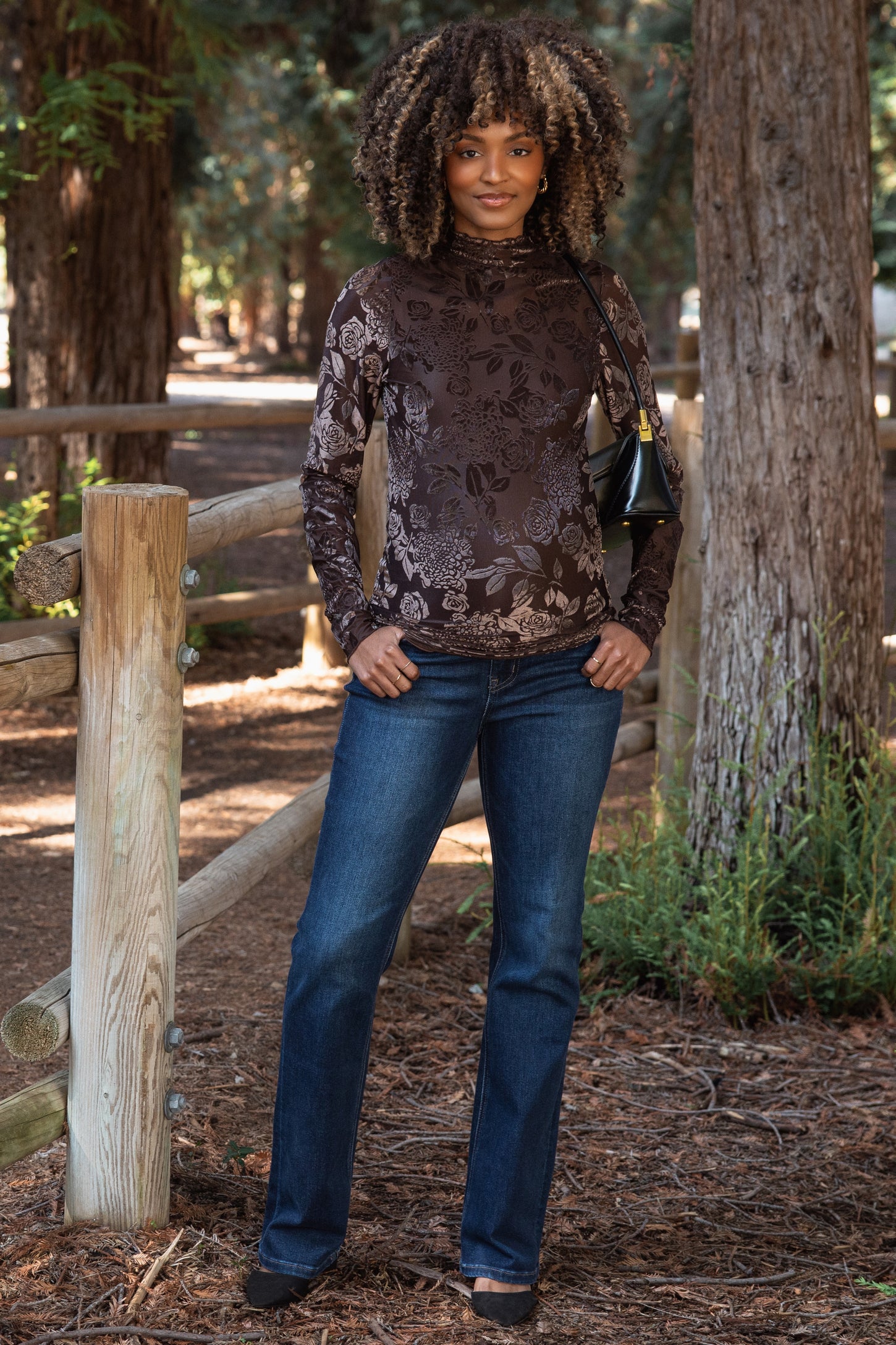 woman wearing a brown floral maternity top and dark jeans, standing outdoors among trees