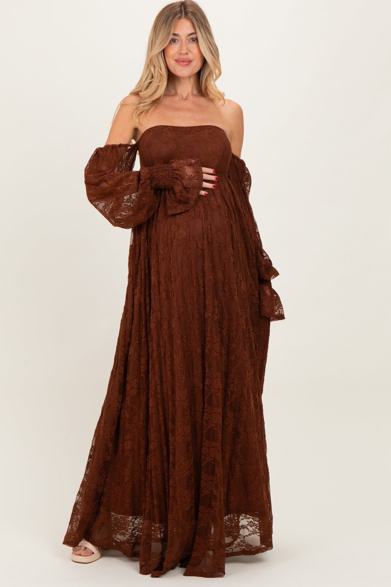 woman wearing a brown lace off-the-shoulder maxi maternity dress, cradling her baby bump