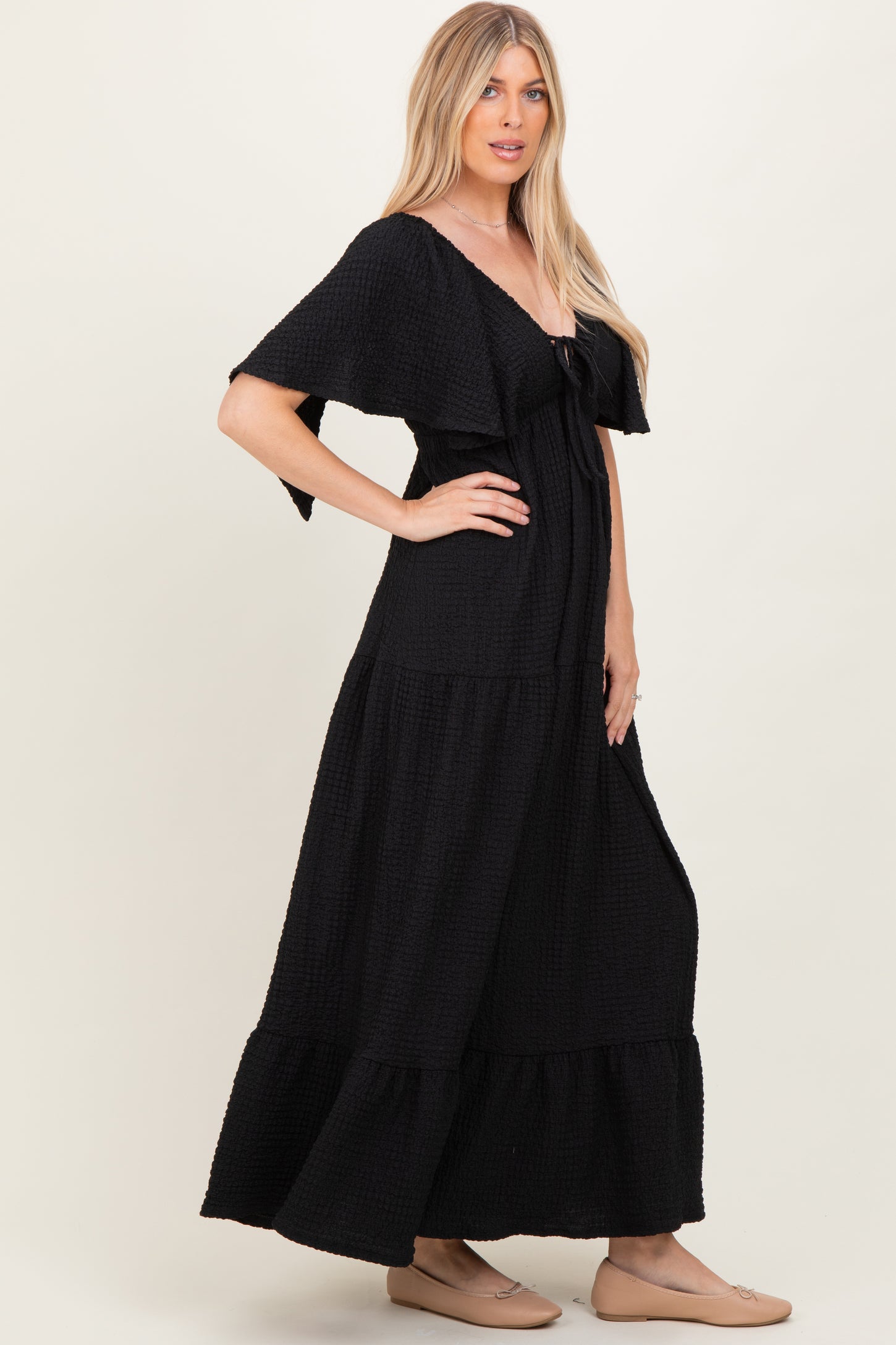 woman wearing a black maxi dress with a tiered design and flutter sleeves, standing in profile against a neutral background