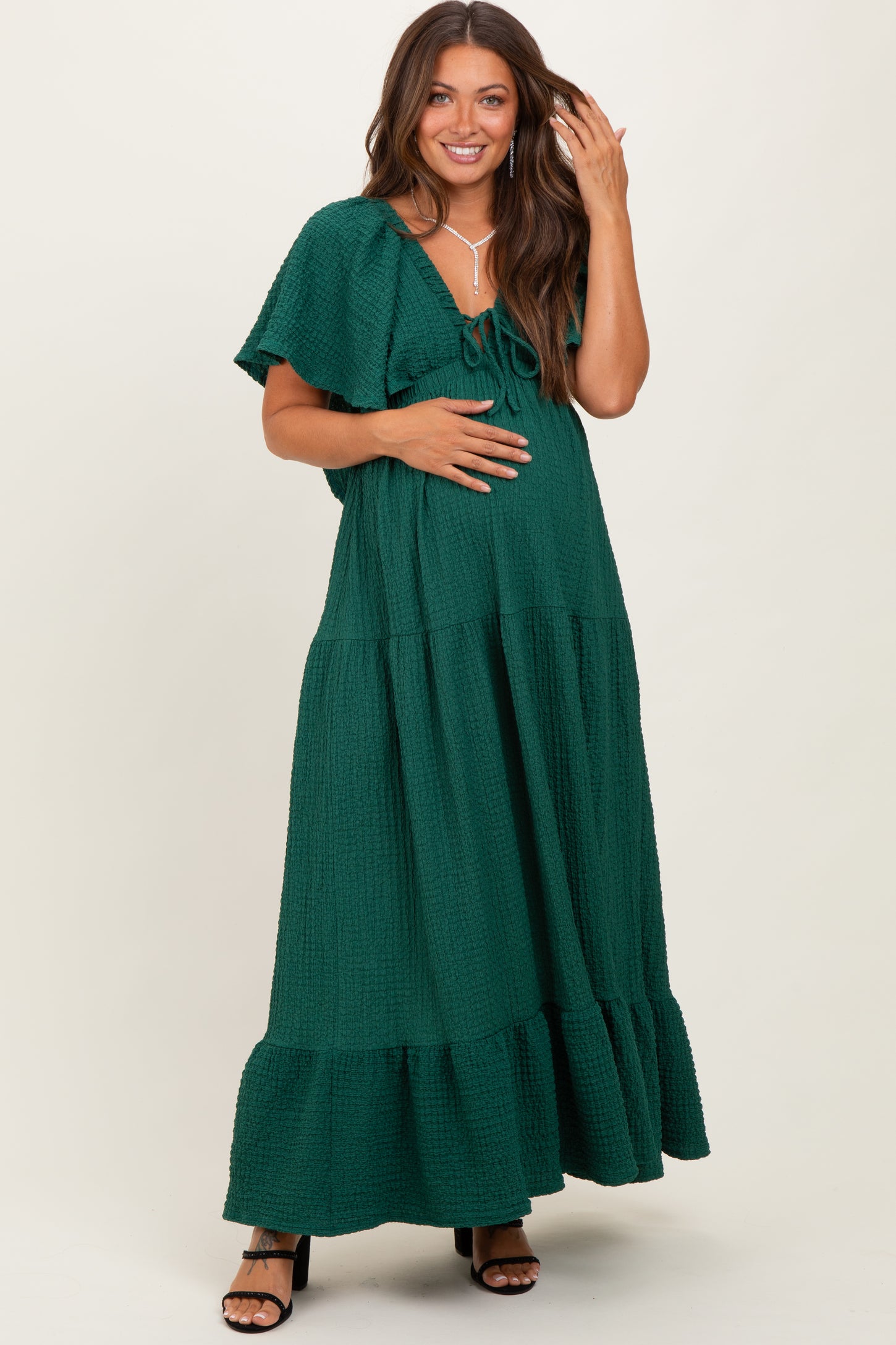 woman wearing a green tiered maternity maxi dress with short sleeves and a tie detail, holding her baby bump