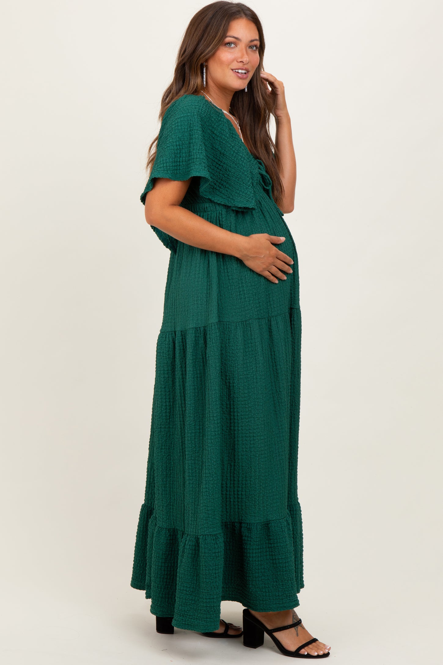 woman wearing a dark green maternity maxi dress with tiered ruffles and a tie front, gently cradling her baby bump