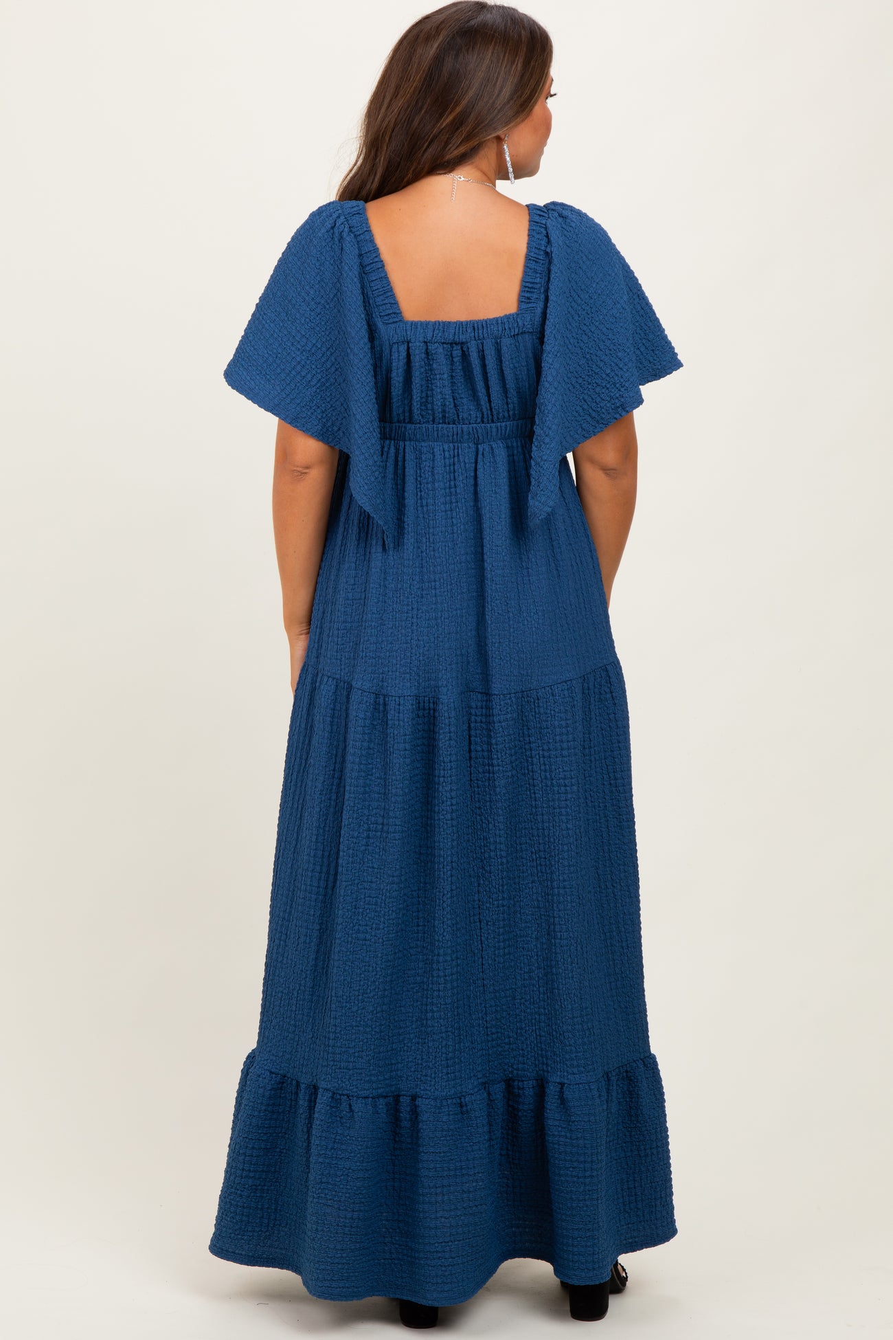 woman wearing a navy blue flutter sleeve maternity maxi dress with a tiered skirt and square back detail