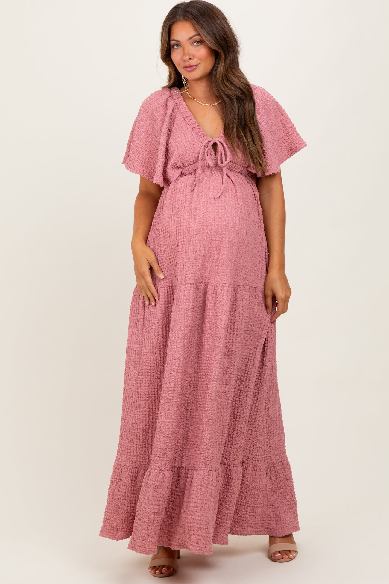 woman wearing a pink maternity maxi dress with short flutter sleeves and a tie detail at the waist, showcasing her baby bump