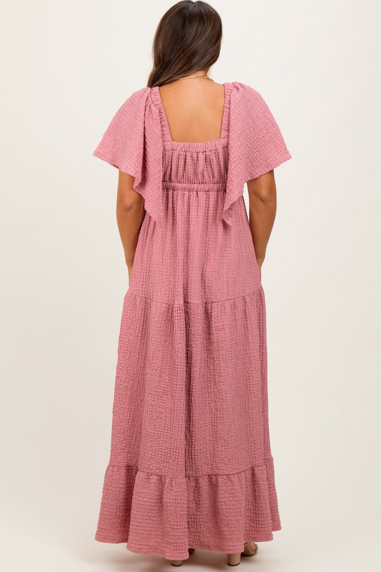 woman wearing a light pink maternity maxi dress with flutter sleeves and a tiered skirt from the back