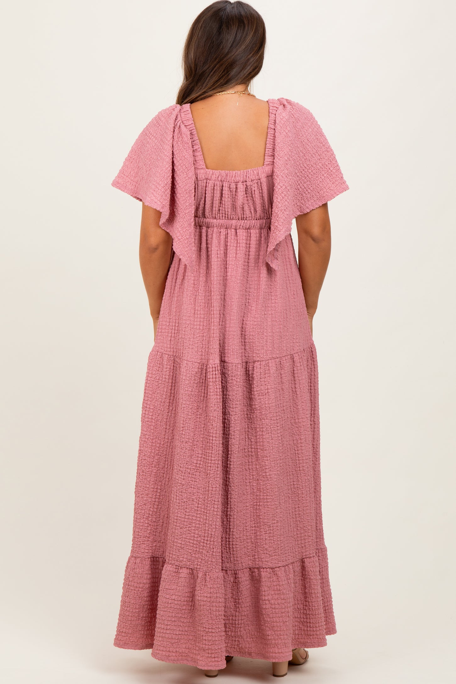 woman wearing a light pink maternity maxi dress with flutter sleeves and a tiered skirt from the back