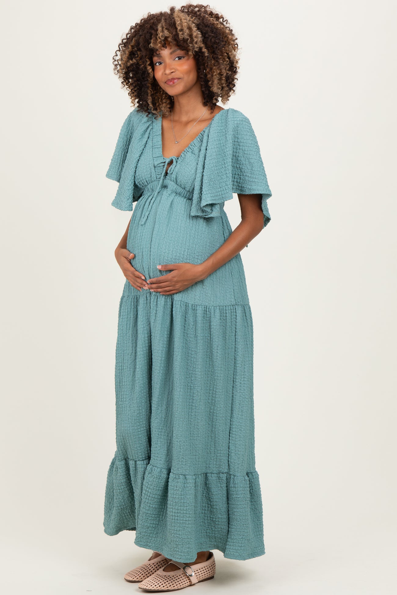 woman wearing a teal maternity flowy maxi dress with short sleeves and a ruffled hem, holding her baby bump