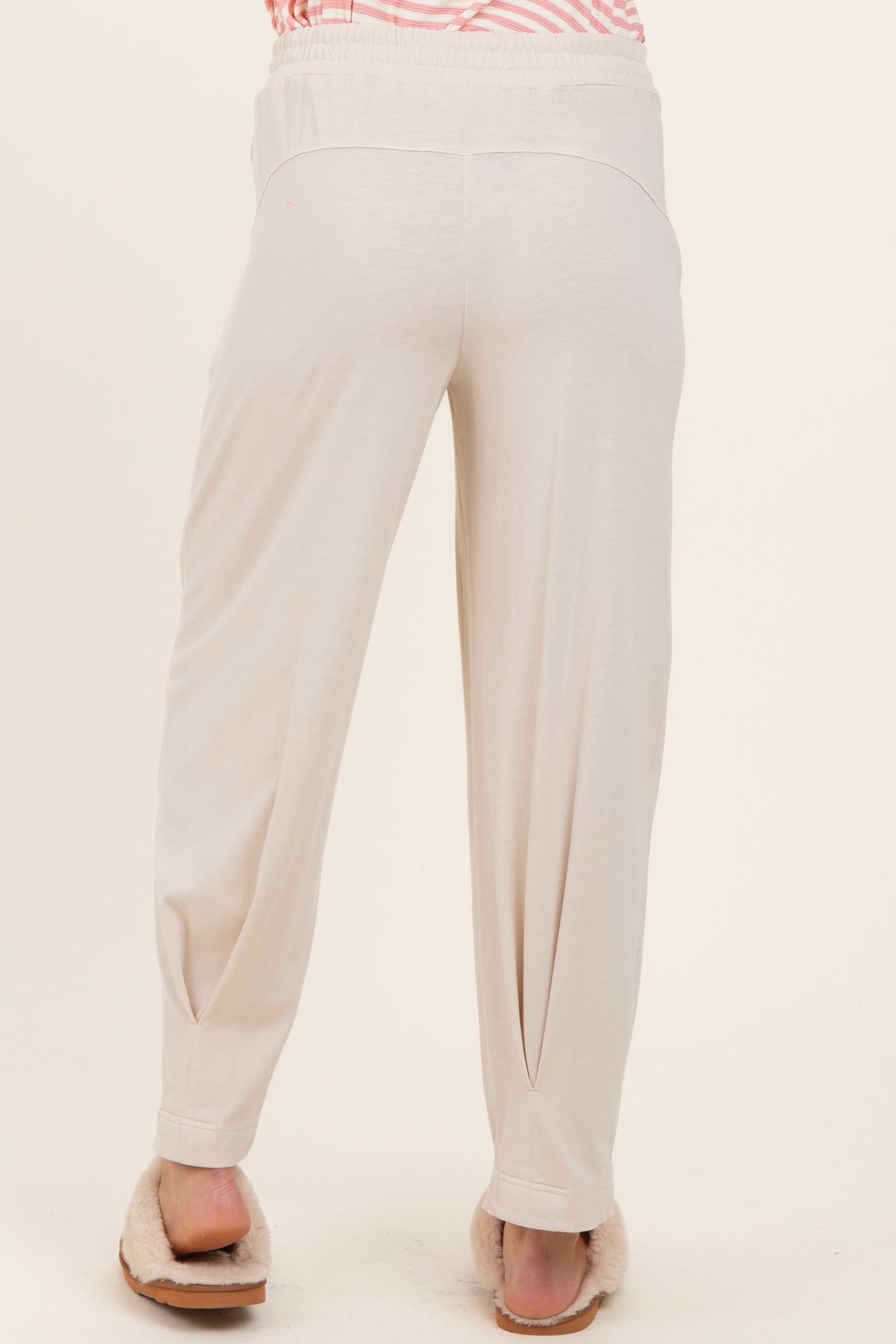 woman wearing cream-colored maternity pants with a relaxed fit, showing the back view and paired with cozy slippers