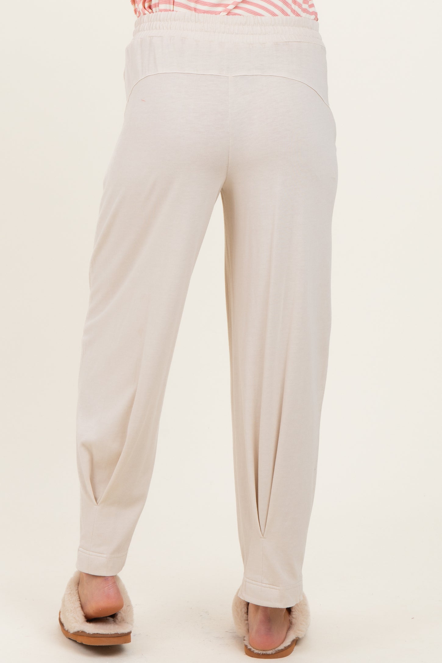 woman wearing cream-colored maternity pants with a relaxed fit, showing the back view and paired with cozy slippers