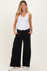 woman wearing a white tank top and black wide-leg maternity pants with pockets, standing with hands in pockets