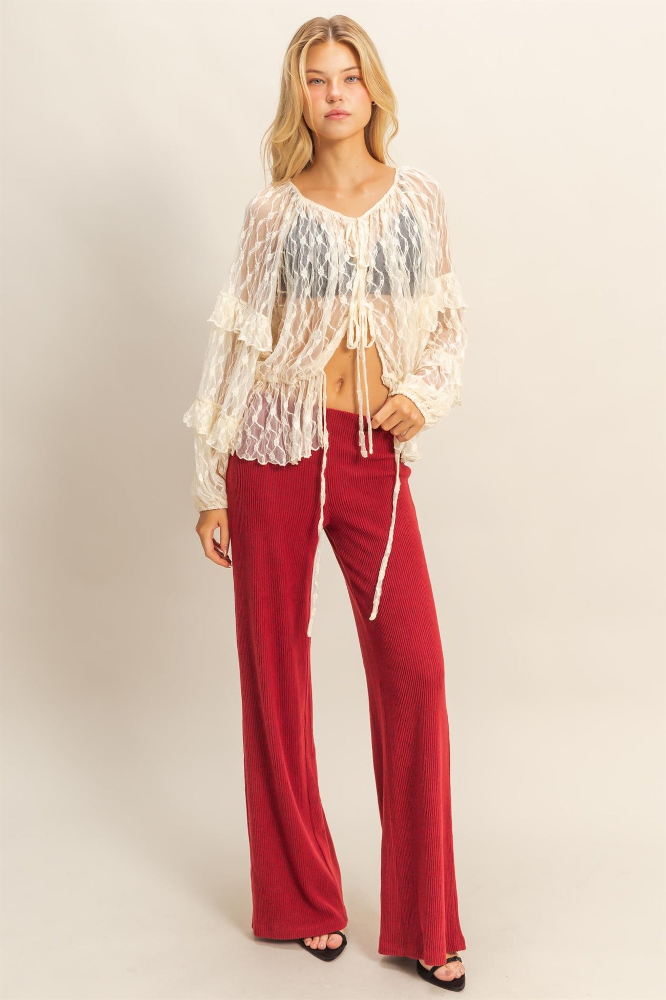 woman wearing a sheer white long sleeve top with ruffles and tie details, paired with red ribbed wide-leg pants