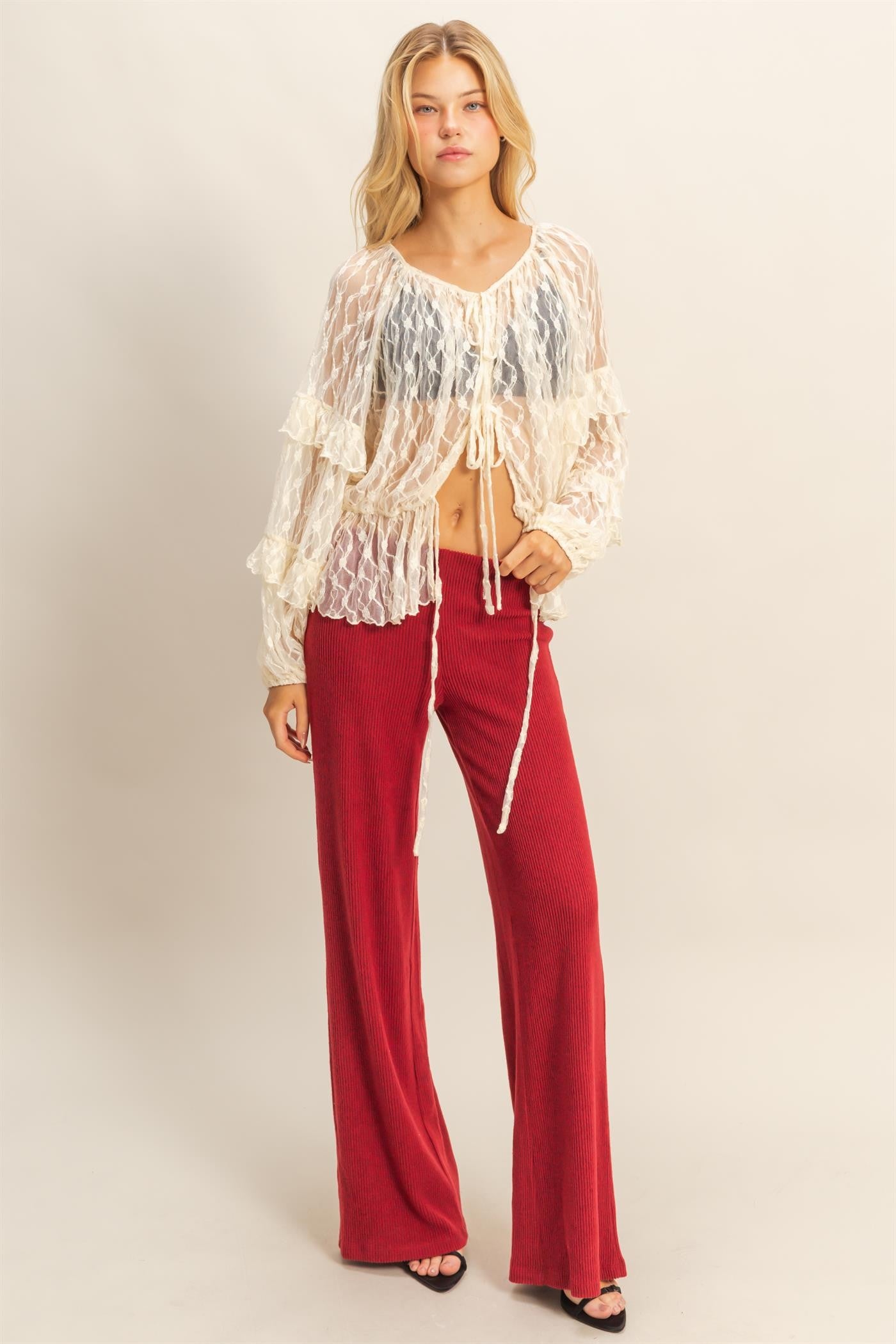 woman wearing a sheer white long sleeve top with ruffles and tie details, paired with red ribbed wide-leg pants