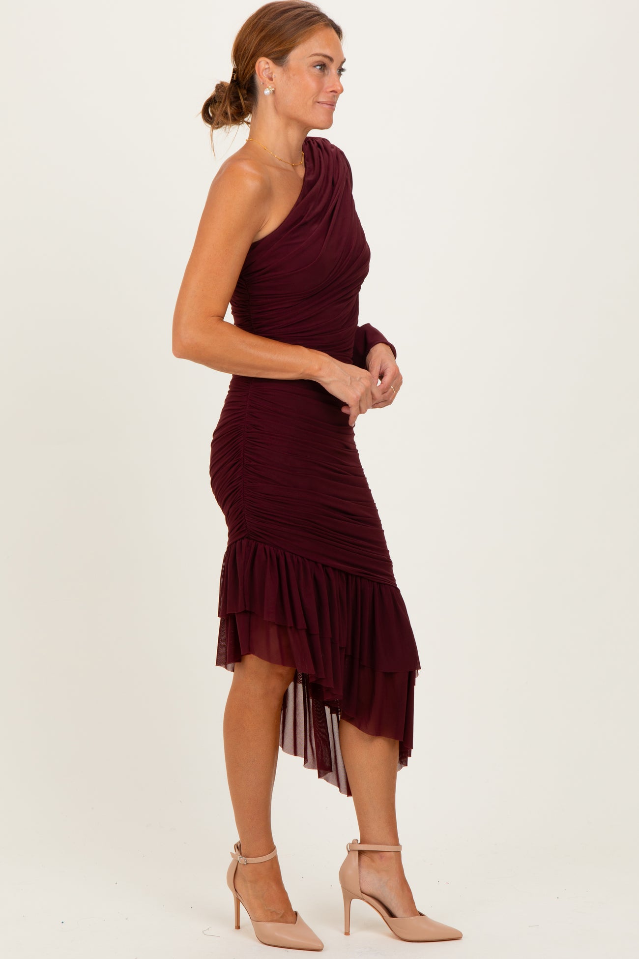 woman wearing a burgundy one-shoulder bodycon dress with ruched detail and a ruffled hem, paired with nude heels