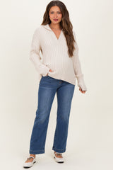 woman wearing a light beige maternity knit sweater with a v-neck and long sleeves, paired with blue flared jeans and white shoes