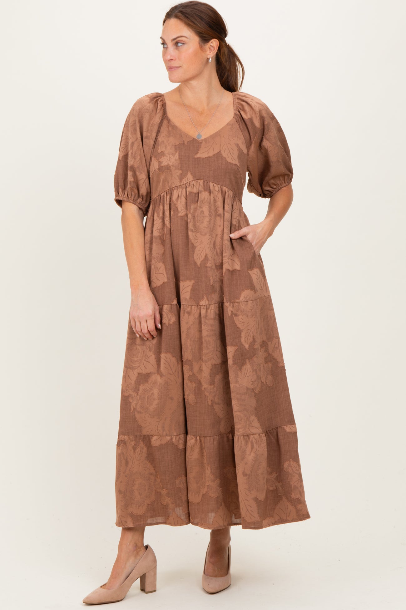 woman wearing a brown floral maxi dress with puff sleeves and a tiered skirt, standing with one hand in the pocket