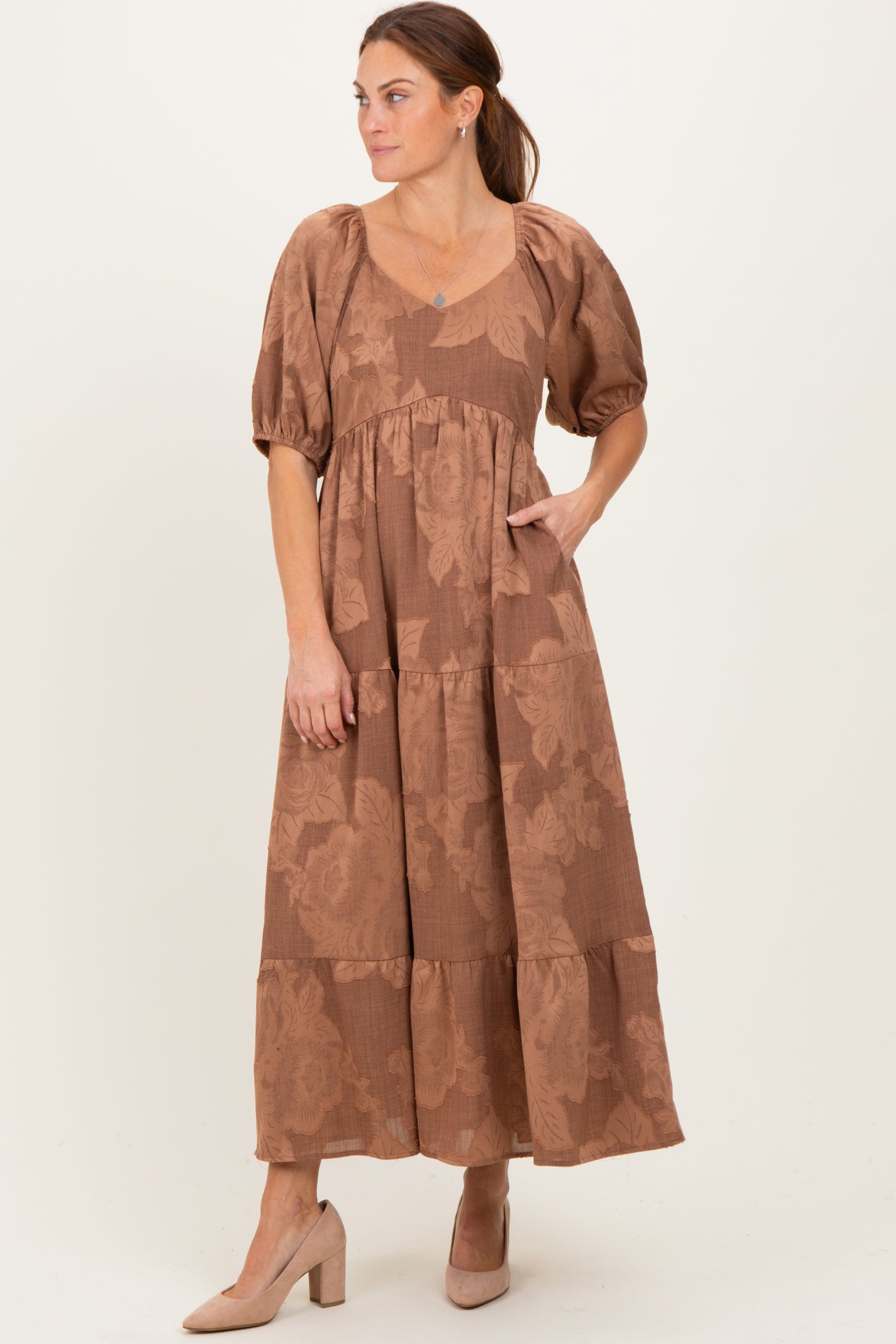 woman wearing a brown floral maxi dress with puff sleeves and a tiered skirt, standing with one hand in the pocket