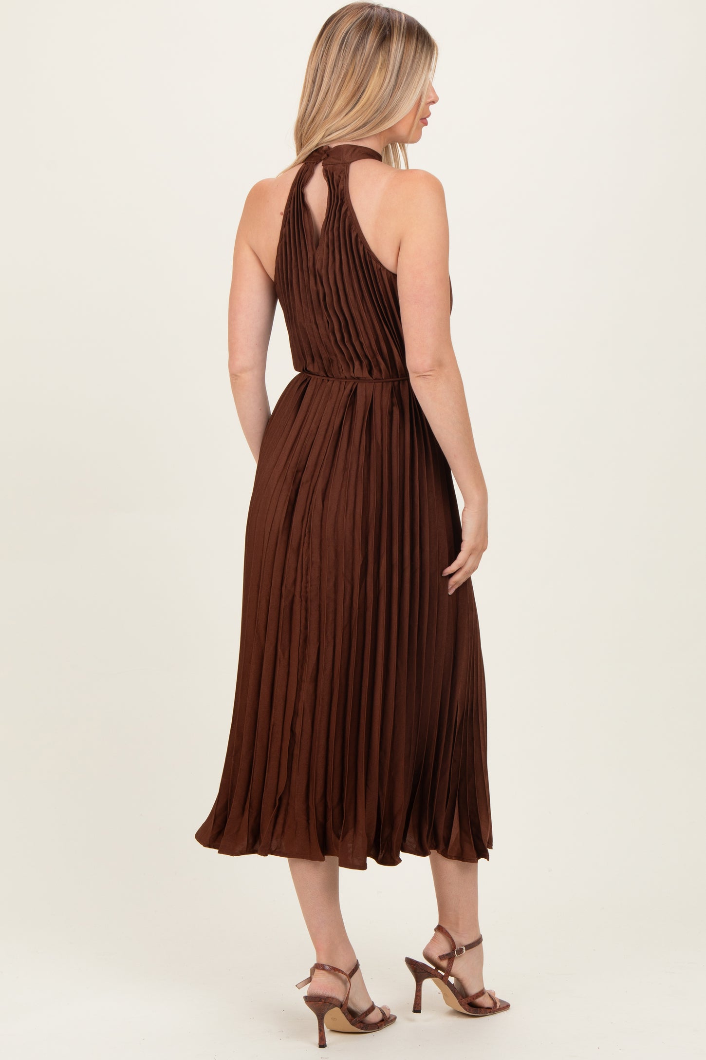 woman wearing a brown pleated midi dress with a halter neck and open back, styled with strappy heels