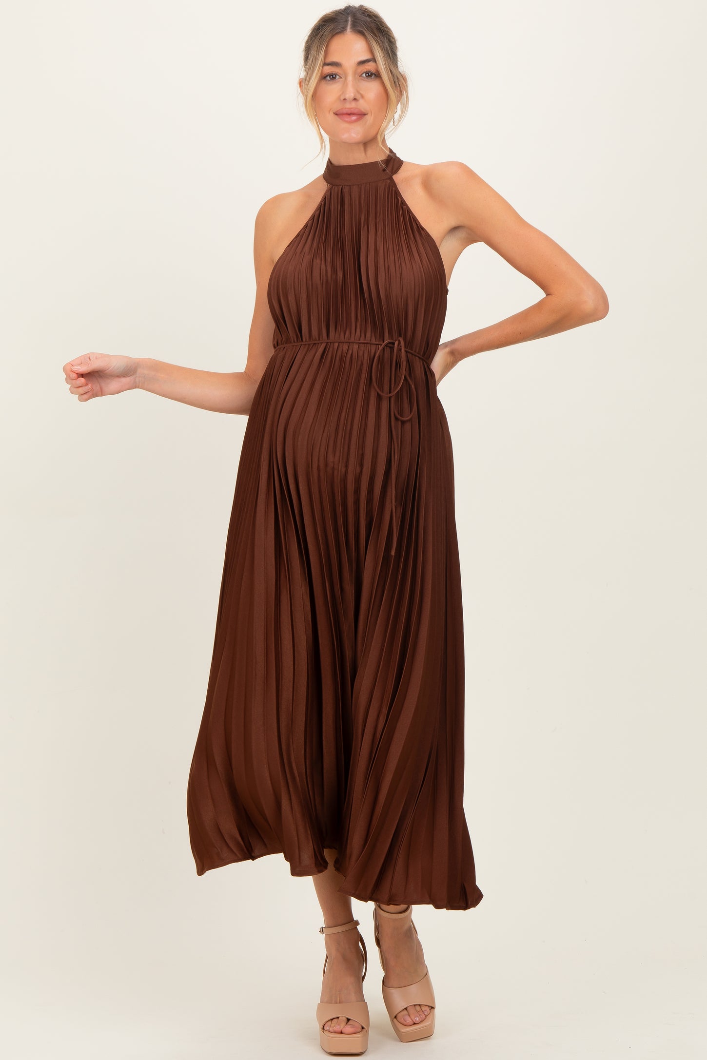 woman wearing a brown pleated maternity dress with a halter neck and a tied waist, showcasing her baby bump