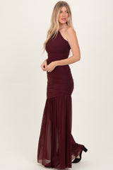 woman wearing a burgundy one-shoulder maxi dress with ruching and a sheer skirt, standing in profile