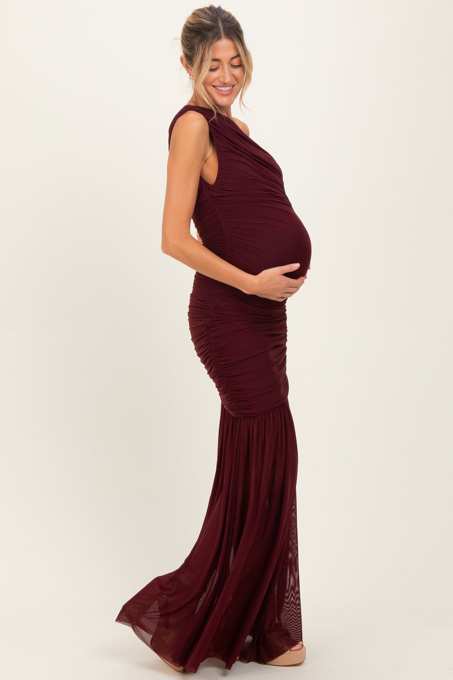 woman wearing a burgundy maternity gown with ruched detailing and a flowing skirt, holding her baby bump