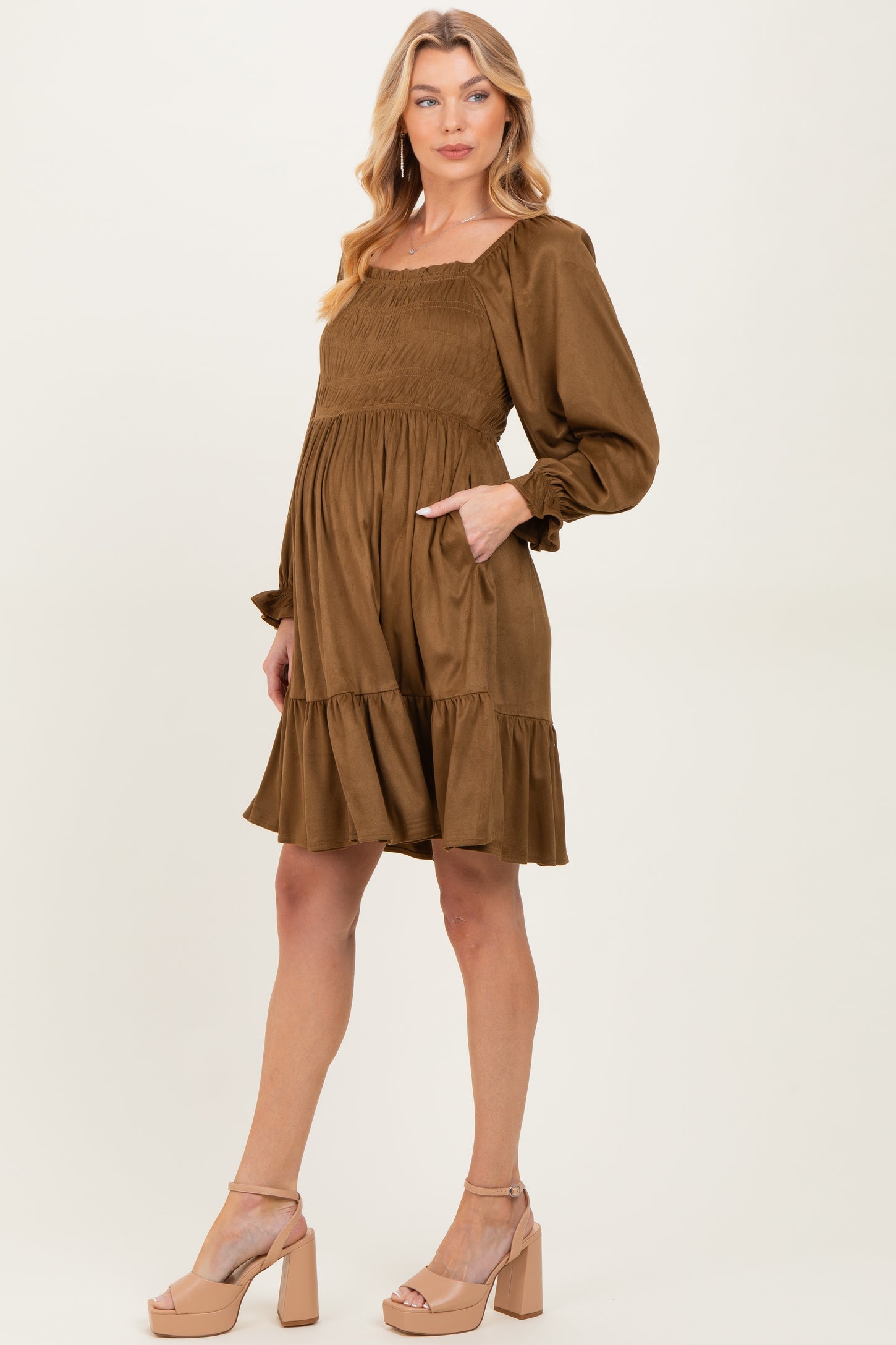 woman wearing a brown maternity dress with a smocked bodice, long sleeves, and a ruffled hem, holding her baby bump