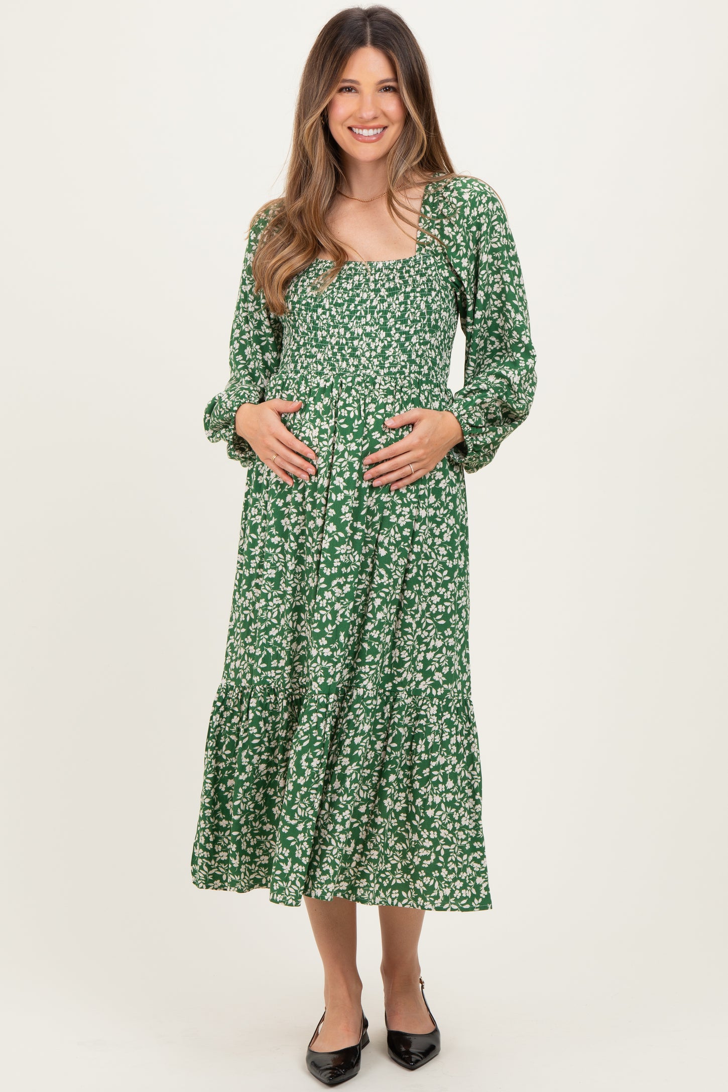 woman wearing a green floral maternity dress with long sleeves and a tiered skirt, holding her baby bump