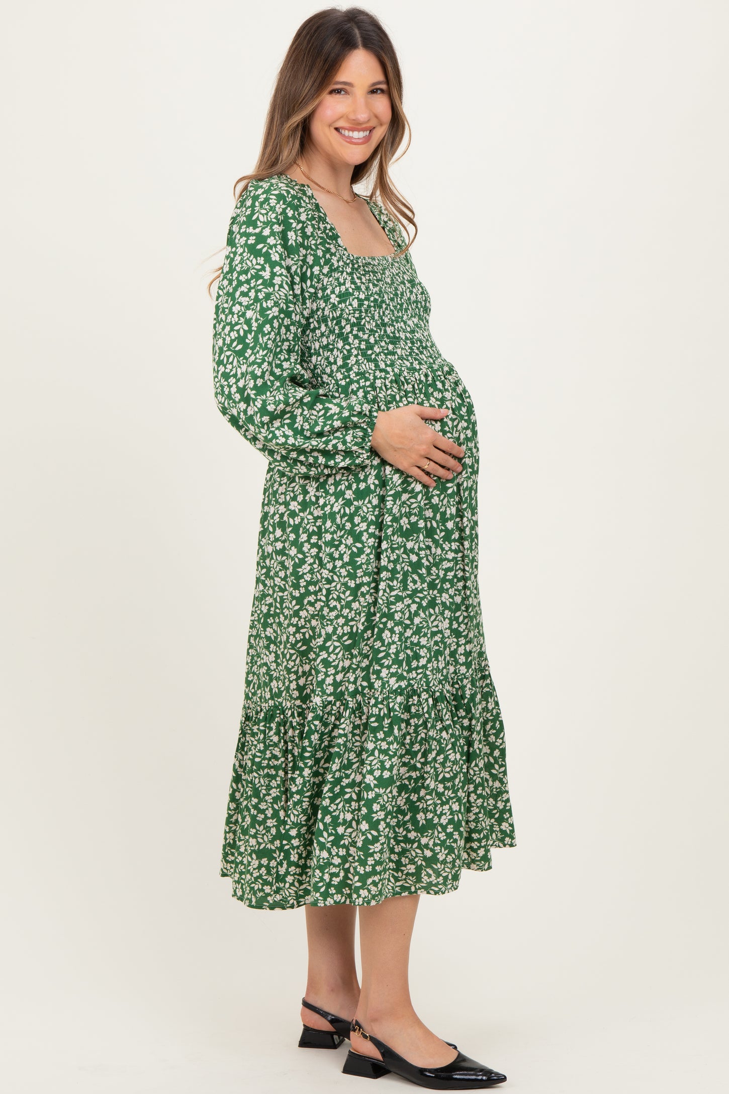 woman wearing a green floral maternity dress with long sleeves and a tiered skirt, holding her baby bump