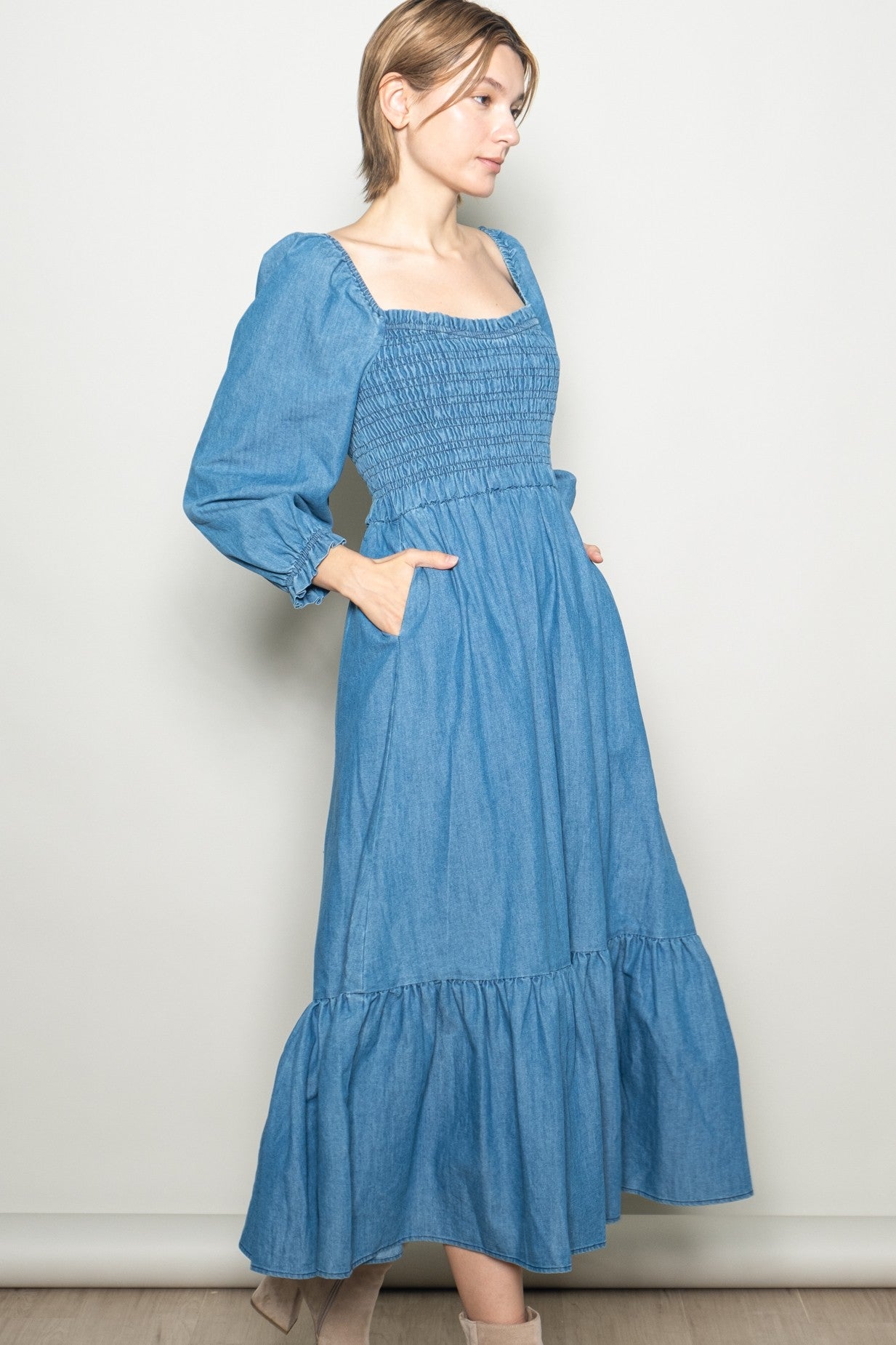 woman wearing a denim blue long sleeve midi dress with a smocked bodice and ruffle hem, standing with hands in pockets