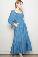 woman wearing a denim blue long sleeve midi dress with a smocked bodice and ruffle hem, standing with hands in pockets