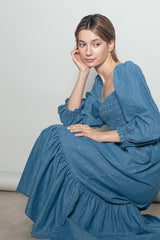 woman wearing a denim blue long sleeve smocked dress with a ruffled hem and sitting gracefully on the floor