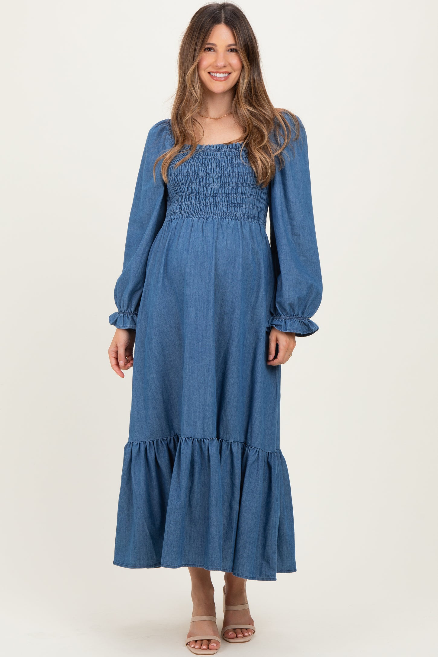 woman wearing a denim maternity maxi dress with smocked bodice and puff sleeves, posing in a neutral background