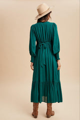 woman wearing a long green tiered dress with balloon sleeves and a tie waist, complemented by a wide-brimmed hat