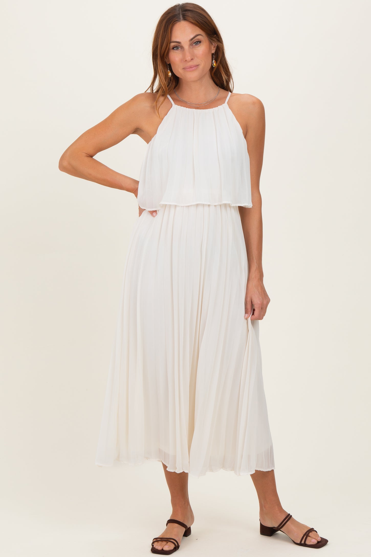 woman wearing a white pleated maxi dress with a halter neckline and layered bodice, standing with one hand on her hip