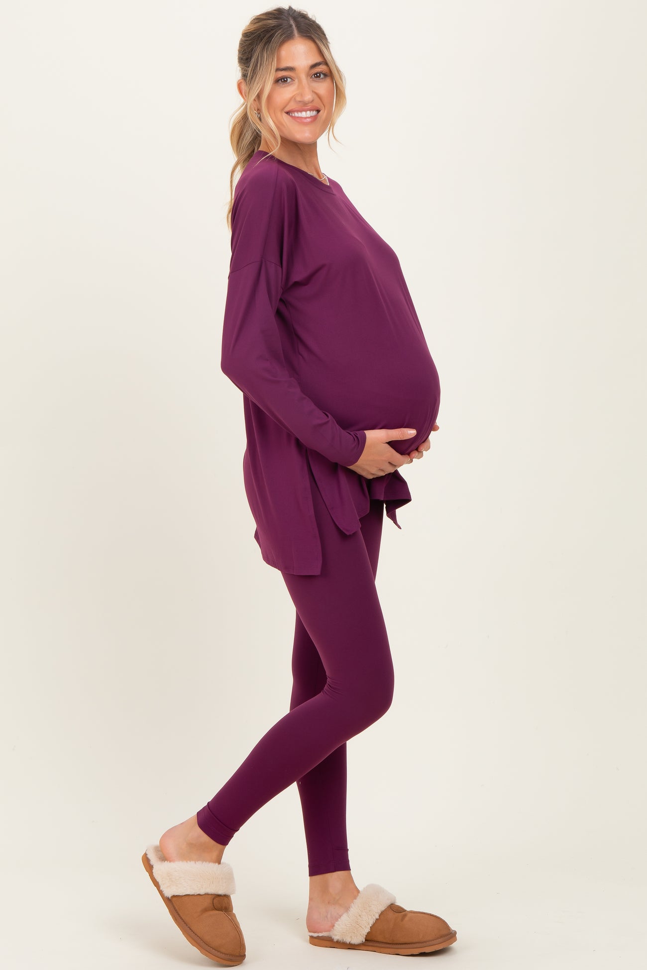 woman wearing a purple long sleeve maternity top and matching leggings, holding her baby bump, styled with cozy slippers