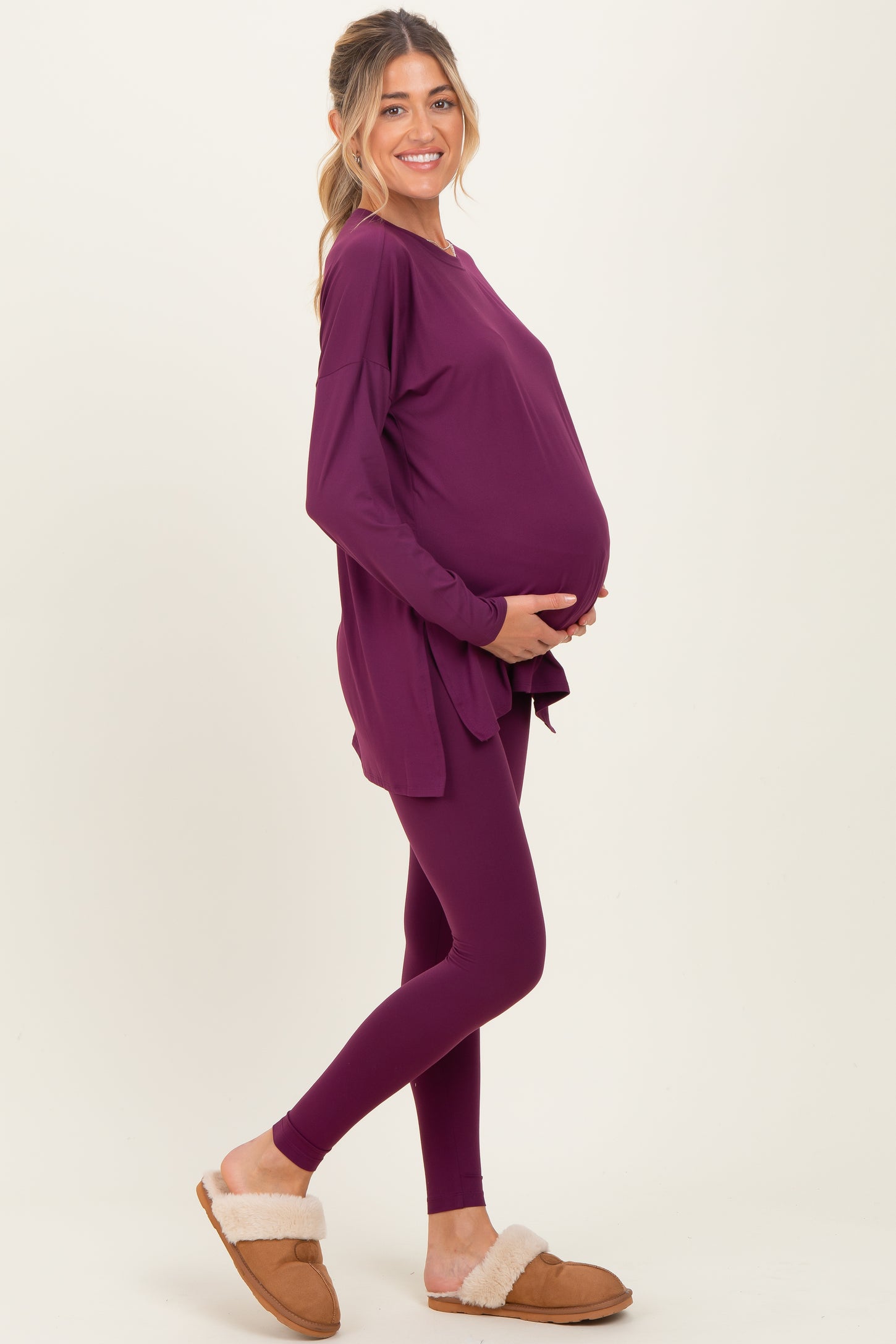 woman wearing a purple long sleeve maternity top and matching leggings, holding her baby bump, styled with cozy slippers