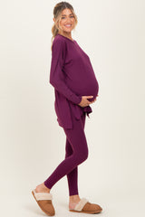 woman wearing a purple long sleeve maternity top and matching leggings, holding her baby bump, styled with cozy slippers