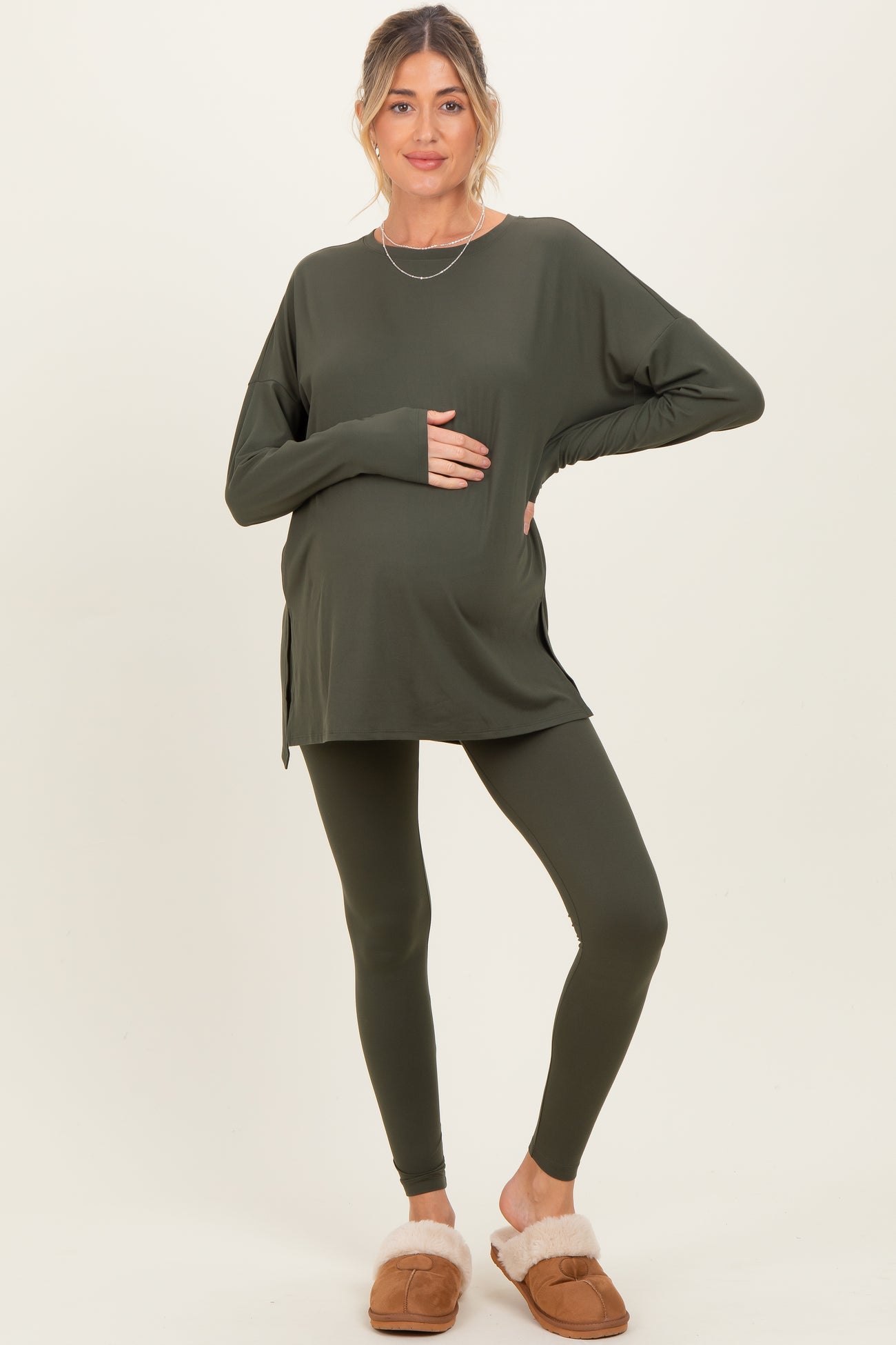 woman wearing a dark green long sleeve maternity top and leggings, holding her baby bump, paired with slippers