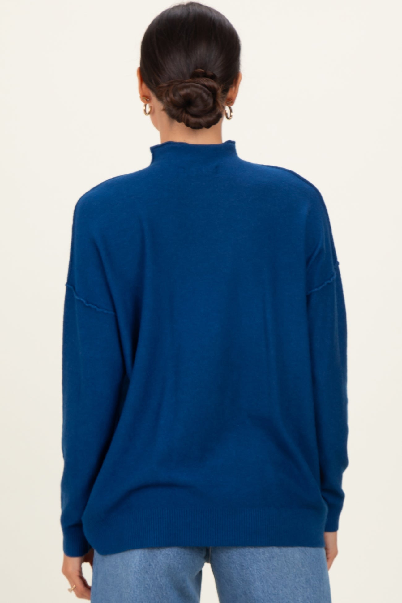 woman wearing a back view of a dark blue oversized sweater with a high neck and relaxed fit, styled with denim jeans