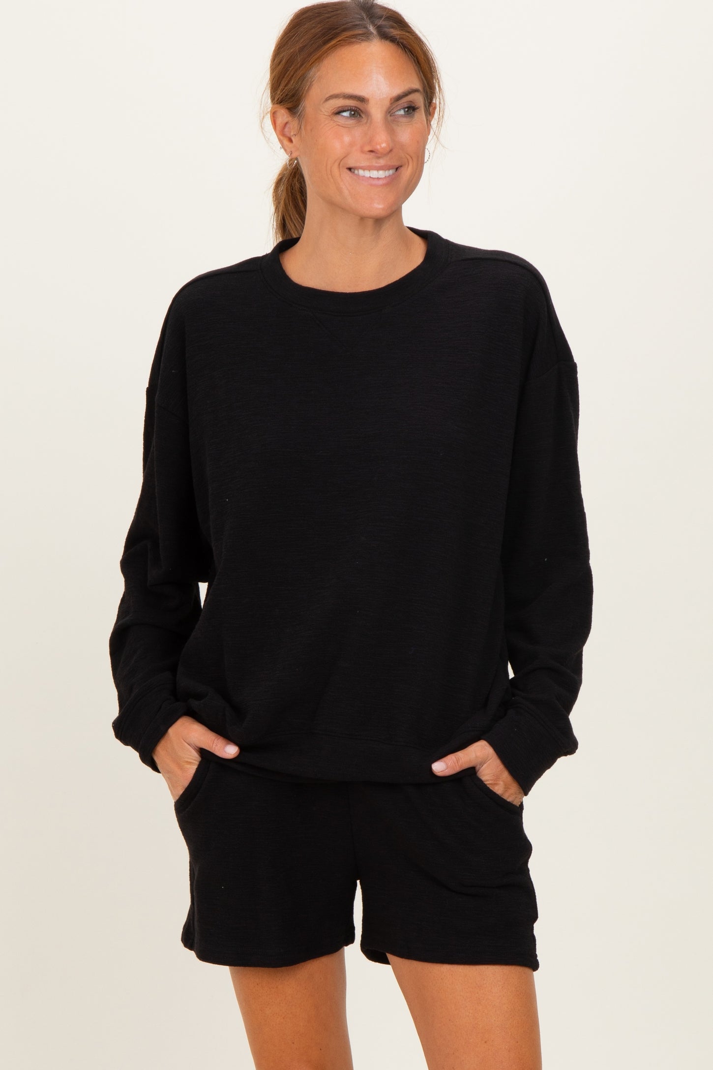 woman wearing a black oversized sweatshirt and matching black shorts, posing with hands in pockets