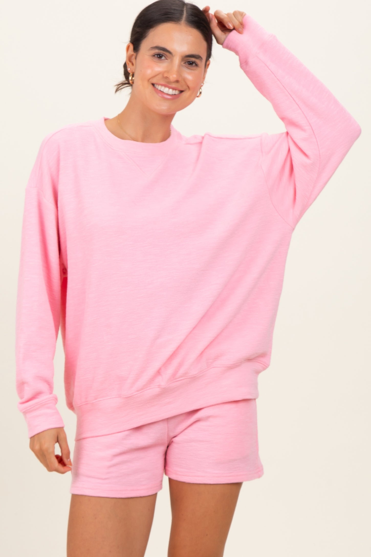 woman wearing a pink oversized sweatshirt and matching pink shorts, smiling and raising one arm