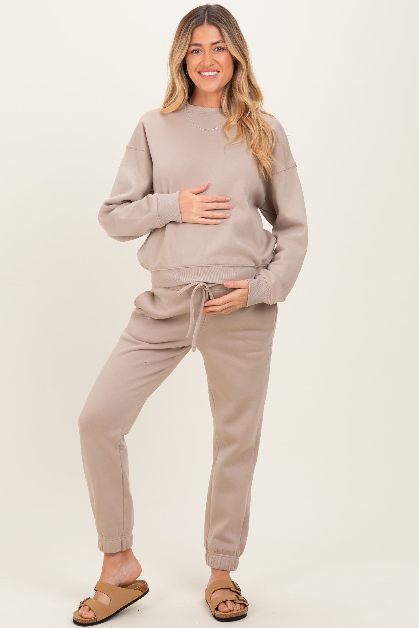 woman wearing a beige maternity sweatshirt and matching joggers, holding her baby bump
