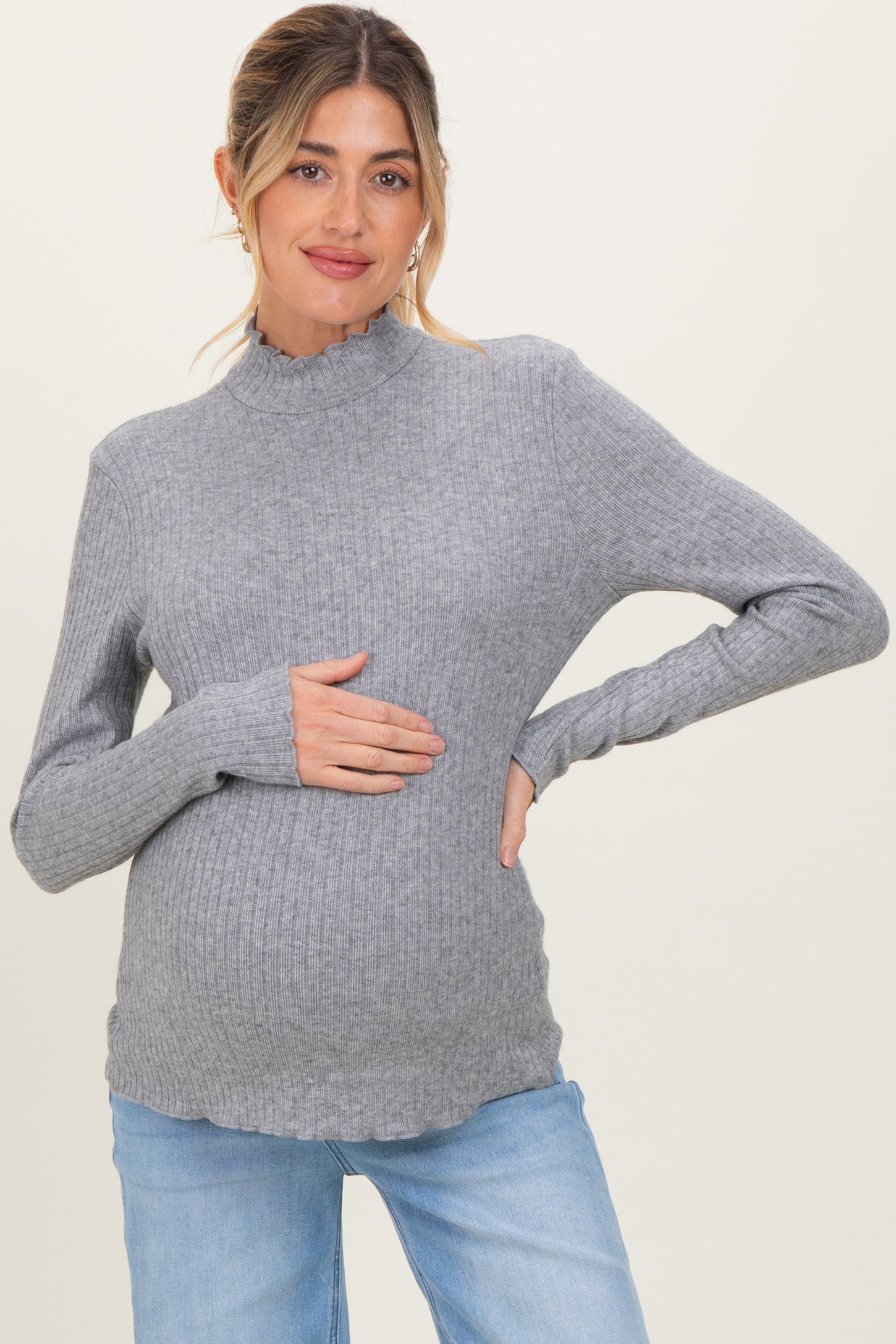 woman wearing a light grey maternity ribbed sweater with ruffled neckline and holding her baby bump