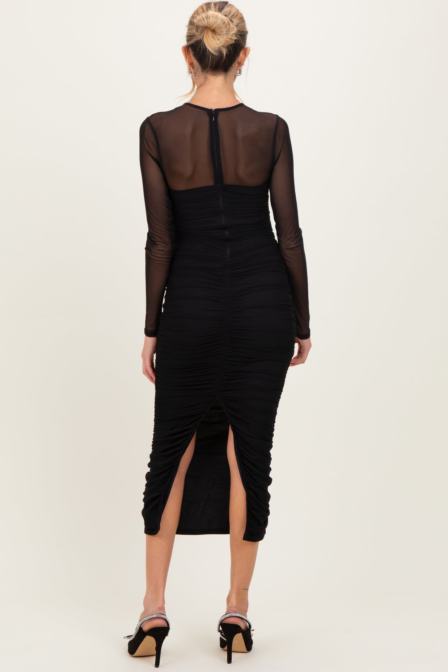 woman wearing a black ruched maternity dress with long sheer sleeves and back slit detail, showing a subtle baby bump
