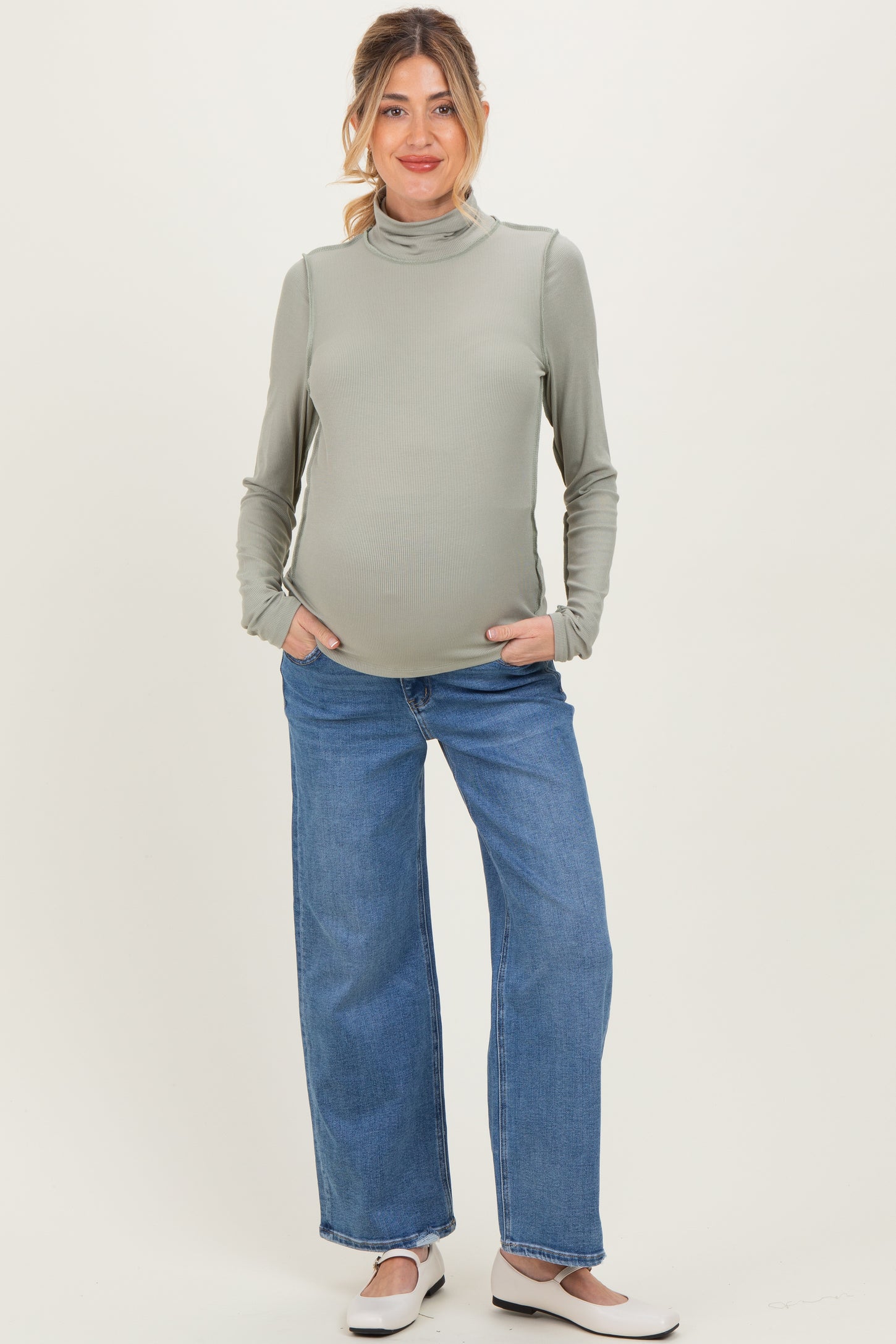 woman wearing a light olive green long sleeve maternity top and blue wide leg jeans, showcasing her baby bump