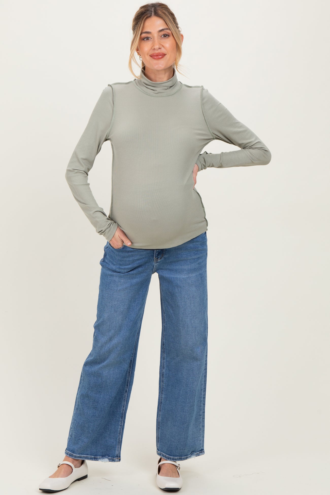woman wearing a soft green long sleeve maternity turtleneck top and blue wide-leg jeans, showing her baby bump