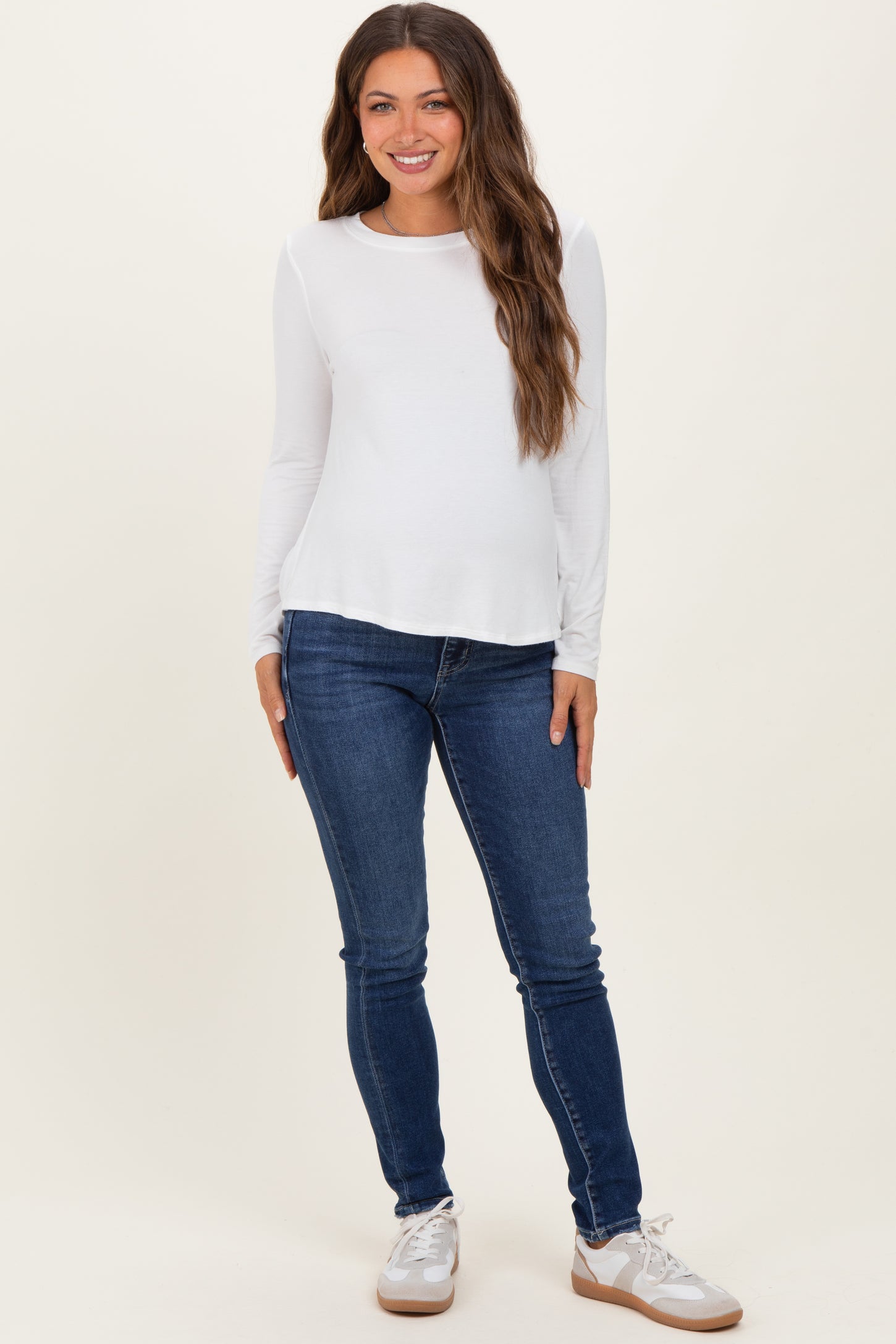 woman wearing a white long sleeve maternity top and blue skinny jeans, showing a baby bump